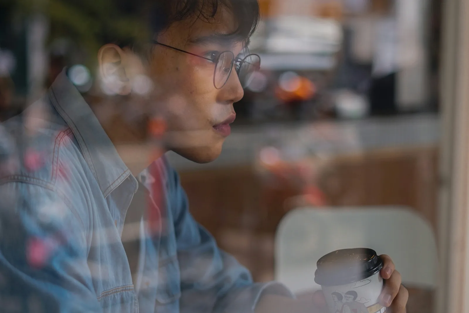 Man in a denim shirt at a cafe window, the street reflected across the glass
